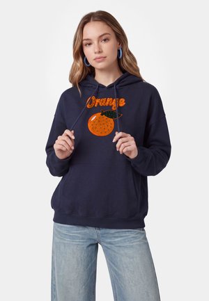 Ung kvinde iført en navy hættetrøje med en orange grafik og tekst, der holder i hættetrådene, kombineret med lyseblå jeans.
