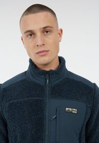 Jonge man met kort haar, gekleed in een marineblauwe fleecejack met een ritssluiting op de borst en een 'PROTEST' logo op de voorkant.