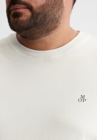 Koszulka z krótkim rękawem, typu crew neck, wykonana z bawełnianej mieszanki w jasnym kremowym kolorze. Na wysokości dekoltu znajduje się małe, ciemne logo „M OP”. Miękka tekstura.
