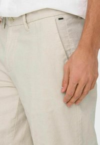 Beige linen pants with a straight cut, featuring side pockets, a button closure, and a subtle black tag detail on the waistband.