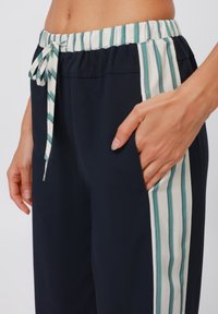 Navyblaue Hose mit einer weißen und türkis gestreiften Seitenpartie, elastischem Bund und Kordelzug. Eine Hand in der Seitentasche.