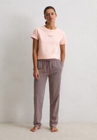 Een vrouw staat blootsvoets en draagt een lichtroze shirt met korte mouwen en een patroon op ruime, loszittende broek met trekkoord tegen een witte muur.