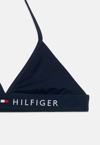 Τοπ μαγιό τρίγωνο σε ναυτικό μπλε, κατασκευασμένο από λείο ύφασμα, με ελαστική ταινία με το λογότυπο "HILFIGER" και ένα κόκκινο και λευκό σήμα σημαίας.
