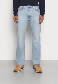 Person wearing light blue straight-leg jeans, brown New Balance sneakers, and a dark blue ribbed sweater standing on a grey floor.