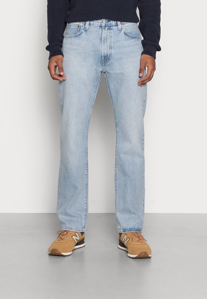 Person wearing light blue straight-leg jeans, brown New Balance sneakers, and a dark blue ribbed sweater standing on a grey floor.