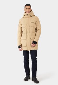 Beige geïsoleerde parka met een capuchon, uitgerust met meerdere voorkwesten en een zachte textuur, gecombineerd met donkere jeans en zwarte laarzen.