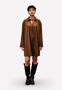 Frau mit kurzen lockigen Haaren, die ein lockeres braunes Langarmkleid und schwarze knielange Stiefel trägt, steht vor einem einfarbigen Hintergrund.