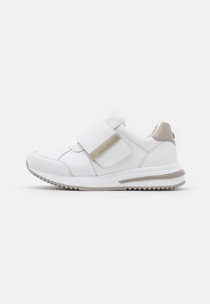 Biały sneaker z beżowymi akcentami, wyposażony w szeroki pasek na rzep i teksturowaną podeszwę zapewniającą przyczepność, widoczny z boku na białym tle.