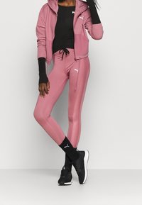 Frau mit pinken Puma-Leggings und -Jacke, schwarzem Top, schwarzen Socken und schwarzen Puma-Sneakern, stehend vor einem schlichten Hintergrund.