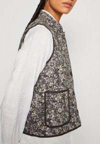 Vesta quilttad med ett blommönster i blått och kräm på en mörkbrun bakgrund. Inkluderar en ståkrage och detalj med framfickor.