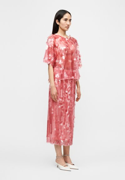 Ροζ μπλούζα με παγιέτες, σχεδίαση με στρώσεις και κοντά μανίκια, σεταρισμένη με μακρύ φούστα ίδιου θέματος, με διαφανή υφή. Λευκά παπούτσια με τακούνι.