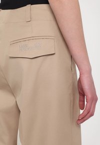 Pantalon beige avec rabat de poche arrière brodé "BAUM UND PFERDGARTEN", porté avec un haut noir, montrant une main détendue sur le côté.