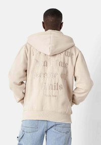 Beige Kapuzenpullover mit einer Kordelzugkapuze und strukturiertem, erhabenem Schriftzug auf der Rückseite. Gerippte Ärmelbündchen und Saum. Weiches Material.