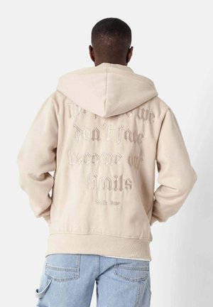 Beige hoodie met een trekkoordcapuchon en een reliëfachtige tekst op de achterkant. Ribgebreide manchetten en zoom. Zacht materiaal.