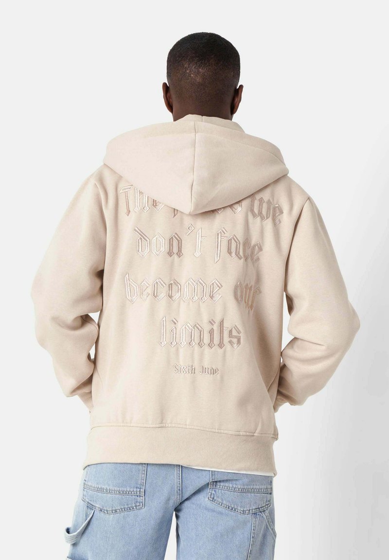 Beige Kapuzenpullover mit einer Kordelzugkapuze und strukturiertem, erhabenem Schriftzug auf der Rückseite. Gerippte Ärmelbündchen und Saum. Weiches Material.