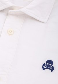 Weißes Polohemd aus Baumwolle mit einem marineblauen, gestickten Totenkopf- und Kruzifix-Logo auf der linken Brust. Verfügt über einen klassischen Kragen und einen Knopfverschluss.