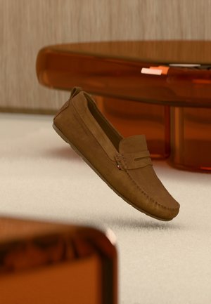 Brauner Wildleder-Loafer mit gesteppten Details, der über einem beigen Teppich schwebt, mit orangenen, durchscheinenden Möbeln im Hintergrund.