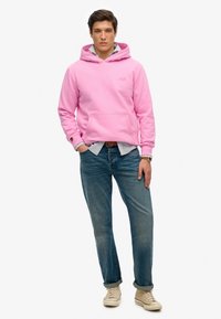 Felpa di cotone rosa con tasca frontale, indossata sopra una camicia a righe blu, abbinata a jeans di denim blu e sneakers color cammello.