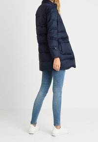 Námořnicky modrý péřový kabát s prošívaným designem, vysokým límcem, bočními kapsami a knoflíkovými detaily, v kombinaci s úzkými modrými džínami a bílými teniskami.