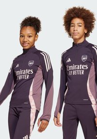 Tenue de sport noir violet avec des accents rose clair, présentant un col montant, des manches longues et un logo. La coupe est ajustée pour la performance.