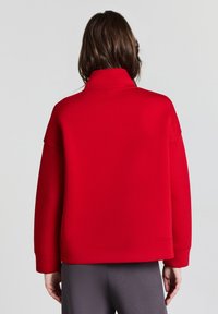 Felpa rossa con colletto alto, vestibilità morbida e maniche lunghe. Texture liscia, polsini e orlo a coste. Indossata con pantaloni sportivi grigi.