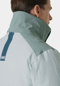 Waterdichte jas met een hoge kraag, lichtblauwe en groene panelen, en een RECCO-logo. Het materiaal lijkt glad en duurzaam te zijn.