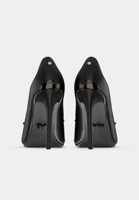 Crne cipele s visokim potpeticama s glatkim sjajem, šiljastim vrhom i vidljivim zlatnim detaljima na potpetici i logotipu marke.