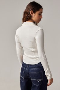 Bela bluzica z dolgimi rokavi s prilegajočim dizajnom in ovratnikom, v kombinaciji z temno modrimi jeans hlačami z kontrastnim šivanjem in žepi.