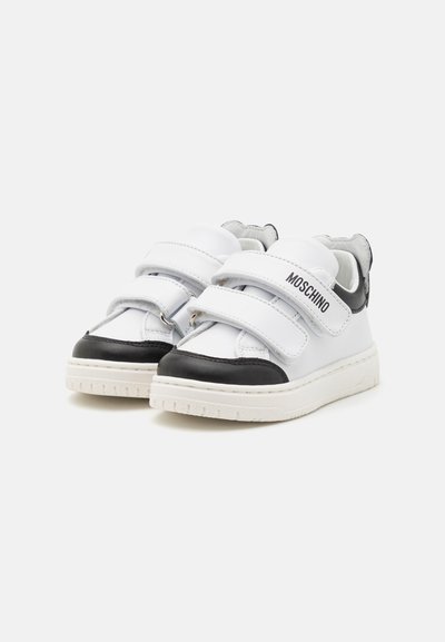 Zapatillas de cuero blancas y negras con tiras de velcro, que presentan una suela blanca texturizada y un logotipo en el lateral. Diseño de punta redondeada.
