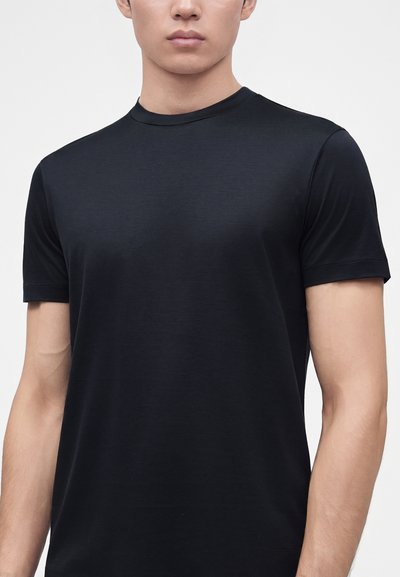 Νέος άνδρας με απλό μαύρο κοντομάνικο T-shirt με στρογγυλή λαιμόκοψη, σε κάδρο από τον λαιμό ως τη μέση του κορμού, σε απλό ανοιχτό φόντο.