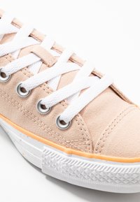 Ljus persika kanvas sneakers med vita snören, silverfärgade öglor och en texturerad vit gummisula med en orange detalj på mellansulan.