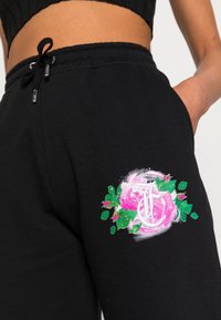Černé tepláky s měkkou texturou mají barevně vyšívaný květinový vzor růžových růží a zelených listů na pravé kapse.