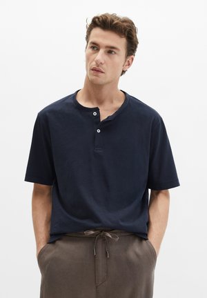 Mężczyzna w granatowej koszulce Henleya z krótkim rękawem i brązowych spodniach z sznurkiem, stojący z rękami w kieszeniach, patrzący lekko na bok.