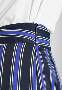 Tmavě modrý materiál s vertikálními pruhy v modré a bílé barvě, s bočním zipem. Hladká textura s definovanými detaily v oblasti pasu.