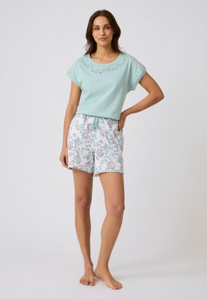 Femme debout pieds nus portant un haut bleu clair à manches courtes avec des découpes et un short blanc à motif floral avec cordon de serrage.