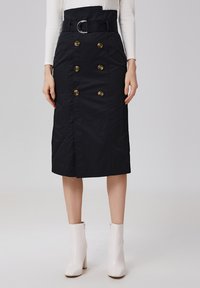 Jupe crayon noire avec une taille ceinturée, ornée de six boutons dorés sur le devant, confectionnée dans un tissu lisse. Associée à des bottines crème.
