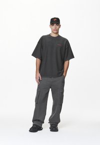 Ung mann står, iført svart caps med oransje logo, oversized mørk grå T-skjorte, løse cargo bukser og svarte sneakers, med hånden i lommen.