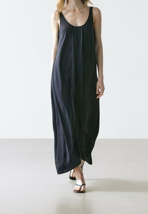 Woman wearing a loose, sleeveless black maxi dress with thin straps, walking barefoot on a smooth, light-colored floor.
