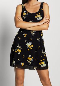 Vestido negro floral con tirantes finos, que presenta flores amarillas y blancas y una textura suave y fluida. El dobladillo llega hasta medio muslo.