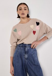 Beige svetr s multibarevnými filcovými aplikacemi ve tvaru srdce, spárovaný s modrými denimovými kalhotami s vysokým pasem, které mají volný střih a skladby.