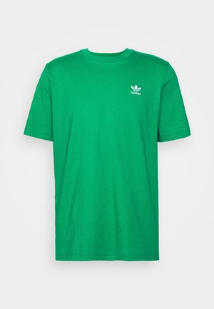 Zielona bawełniana koszulka z okrągłym dekoltem, ciemnoniebieskie dżinsy z podwiniętymi nogawkami oraz szare sneakersy z białymi akcentami. Pomarańczowe obrotowe krzesło w tle.