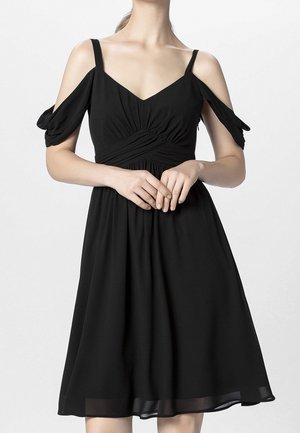 Frau trägt ein schwarzes Cold-Shoulder-Kleid mit dünnen Trägern und gerafften Details an der Taille, steht mit verschränkten Händen.
