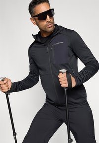 Czarna sportowa kurtka z kieszenią na zamek, w zestawie z czarnymi spodniami i kijkami trekkingowymi; charakteryzuje się dopasowanym krojem i kapturem. Duże ciemne okulary przeciwsłoneczne.