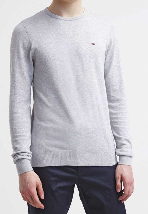 Pull gris clair col rond avec petit logo sur la poitrine, porté par une personne avec les mains détendues le long du corps, associé à un pantalon foncé.