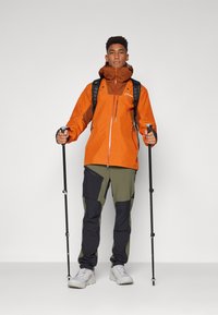 Orange wasserfeste Jacke mit braunen Akzenten; grüne und schwarze Hosen; Trekkingstöcke; graue Wanderschuhe; schwarzer Rucksack.