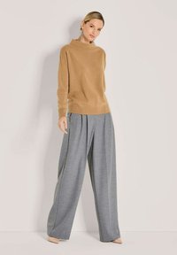 Frau in einem camel-farbenen Rollkragenpullover, weit geschnittenen grauen Hosen, beigen spitzen Schuhen und langen silbernen Ohrringen, die vor einer weißen Wand steht.