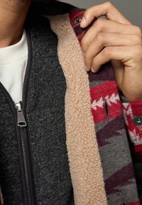 Veste en laine grise avec une doublure en polaire beige, présentant un intérieur à motifs rouges et gris. Fermeture éclair et accents de boutons visibles.