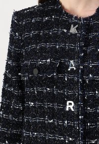 Marinblå tweedjacka med texturerad tyg, med vita och metalliska detaljer, två framfickor och silverbokstäverna "A" och "R" som dekoration.
