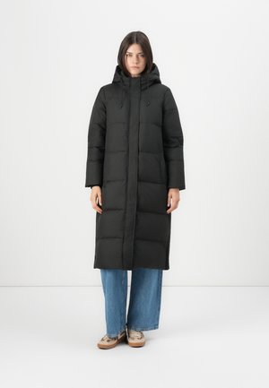 Schwarze Pufferjacke, knielang mit Kapuze, mit einem gesteppten Design, Reißverschluss und Seitentaschen. Getragen über weit geschnittenen blauen Jeans.