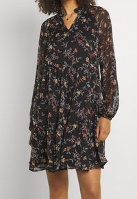 Kvinna iklädd en svart, långärmad blommig klänning med genomskinliga ärmar och en lager-på-lager kjol, stående mot en enkel bakgrund.